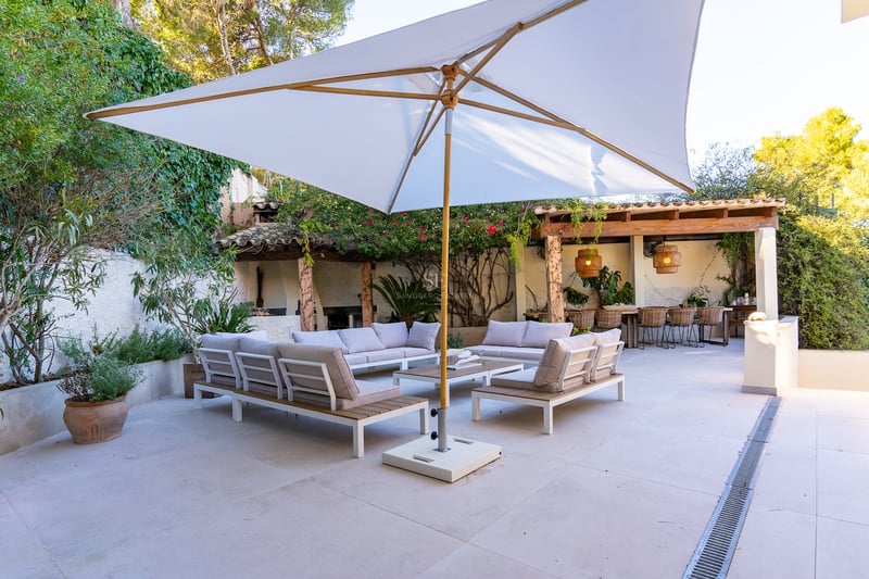 Grande terrasse avec canapés modernes, grand parasol blanc et coin repas sous une pergola entourée de verdure.