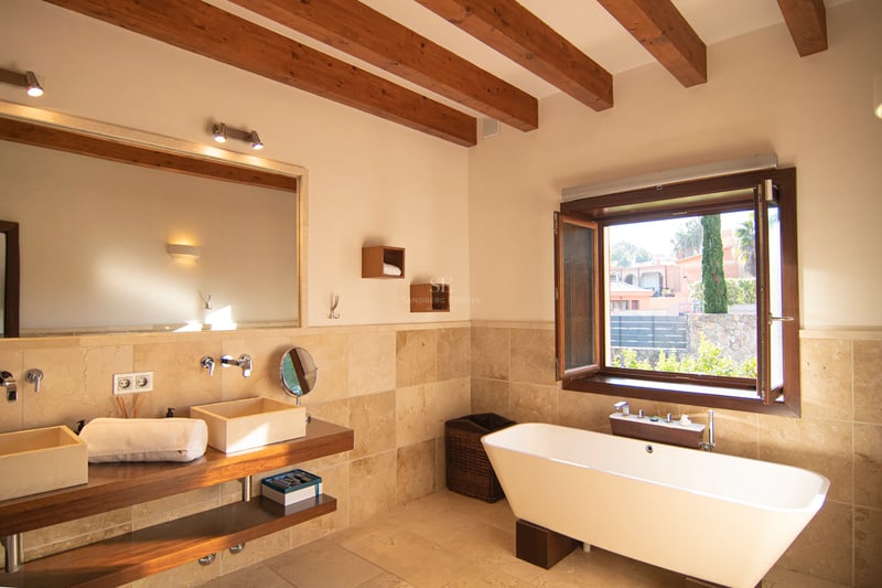 Salle de bains avec poutres apparentes, baignoire îlot, double vasque en pierre et vue sur le jardin.