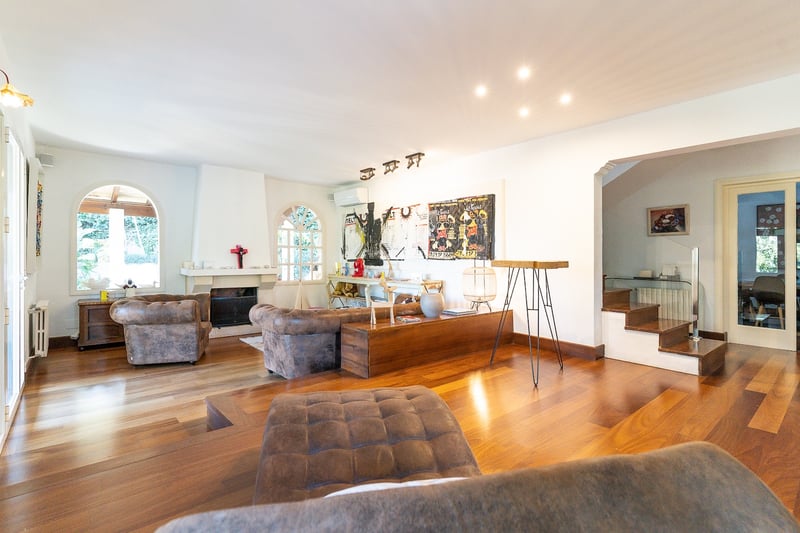 Grand salon avec parquet en bois foncé, fauteuils en cuir, cheminée et fenêtres cintrées sous un éclairage mixte.