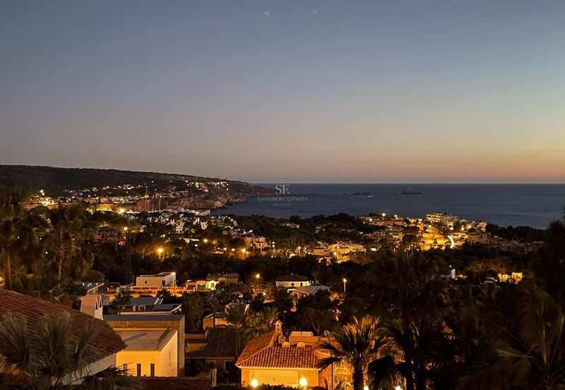 Vue nocturne surélevée d'une côte méditerranéenne avec villas éclairées, palmiers et mer sous un ciel de crépuscule.