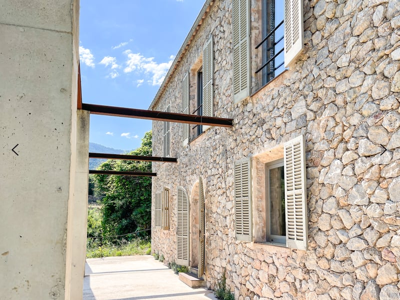 Façade en pierre naturelle d'une villa avec volets vert sauge, poutres en acier industrielles et vue sur la montagne.