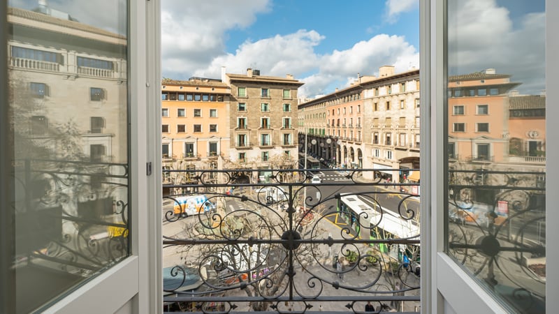 View from a classic balcony with ornate iron railing overlooking a sunlit Mediterranean city square and stone buildings.