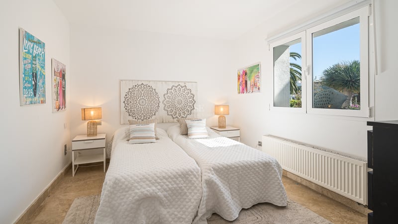 Chambre blanche avec deux lits simples, tête de lit en bois sculpté, lampes en osier et fenêtre sur le jardin.
