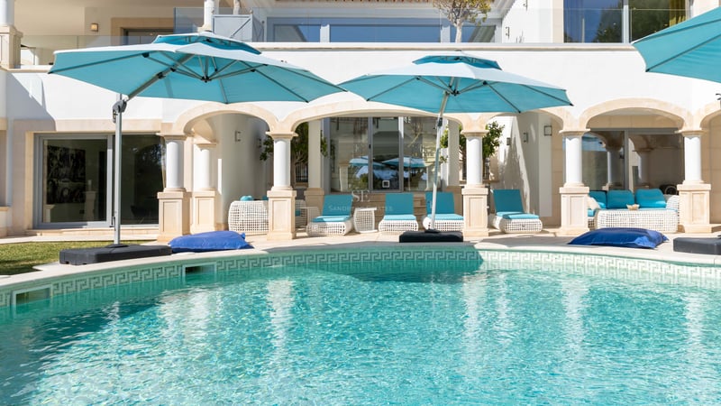 Piscine turquoise avec colonnes blanches classiques et parasols bleu canard sur une terrasse ensoleillée.