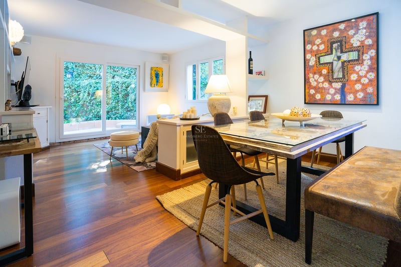 Salle à manger éclairée avec table en verre, chaises modernes et œuvre d'art, vue sur la verdure.
