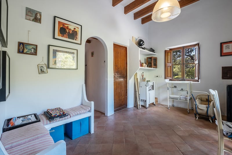 Pièce intérieure avec carrelage en terre cuite, poutres en bois au plafond, murs blancs et banquette intégrée.