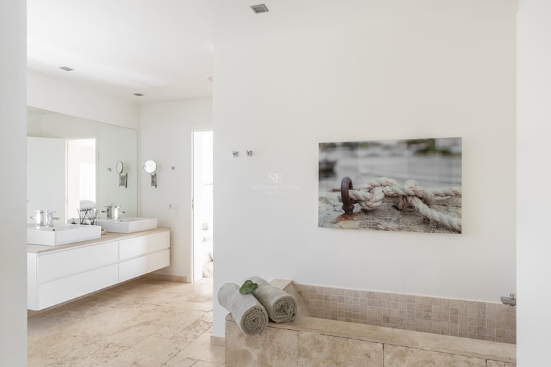 Salle de bains lumineuse avec double vasque suspendue, sol en travertin et baignoire intégrée en pierre.