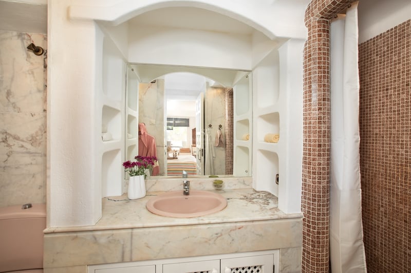 Salle de bain avec vasque rose, plan en marbre, miroir arqué et colonne en mosaïque brune.
