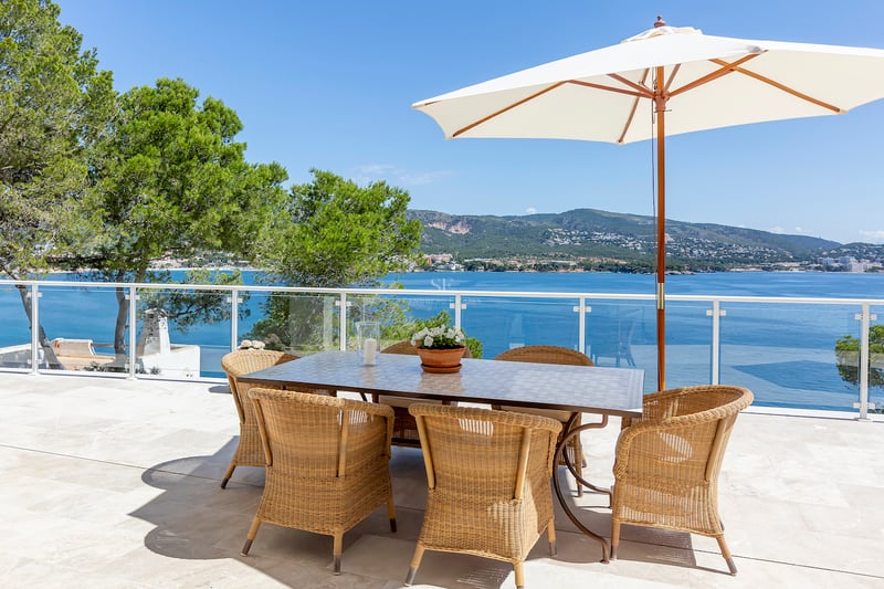 Espace repas extérieur avec chaises en osier et parasol blanc sur terrasse en pierre surplombant mer et montagnes.