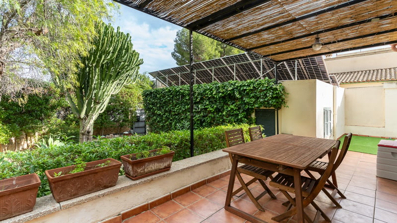 Table à manger en bois sous une pergola en bambou sur une terrasse en terre cuite entourée de plantes et de panneaux solaires.