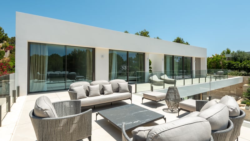 Terrasse de villa moderne avec salon de jardin gris, garde-corps en verre et larges baies vitrées.