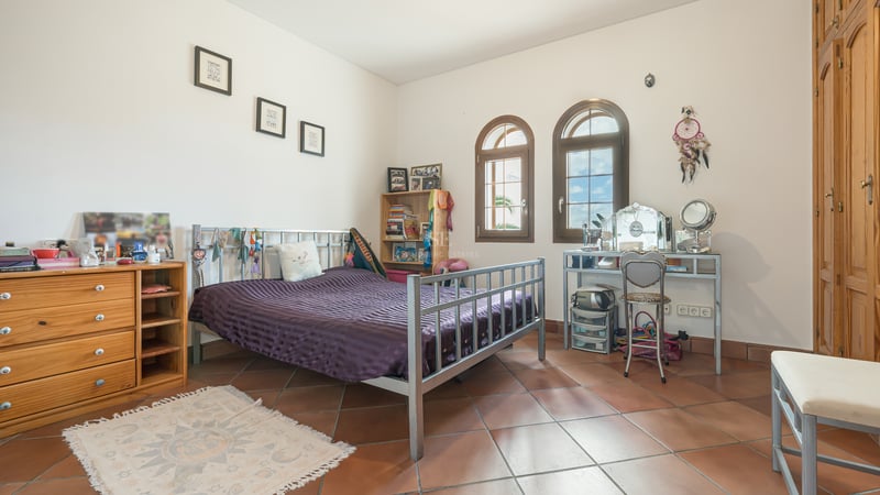 Chambre avec sol en terre cuite, lit en métal, fenêtres cintrées en bois et placards intégrés sous lumière naturelle.