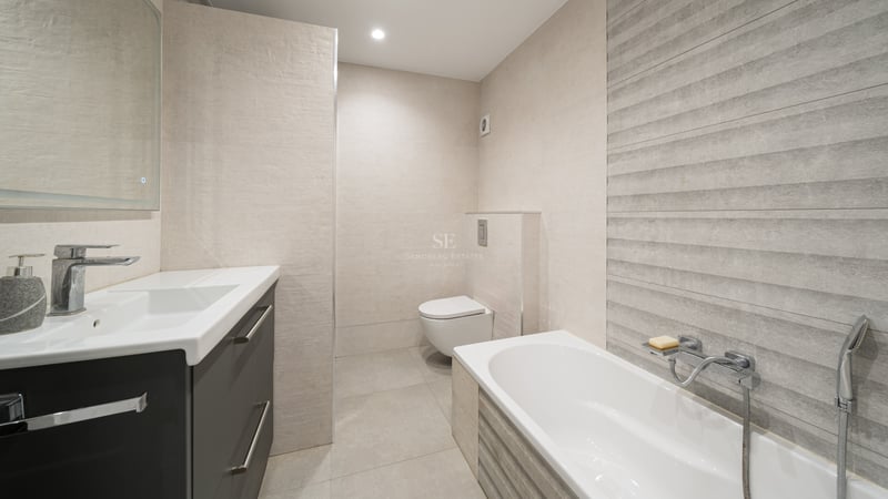 Salle de bain moderne avec baignoire blanche, WC suspendu et meuble gris sur fond de carrelage beige texturé.