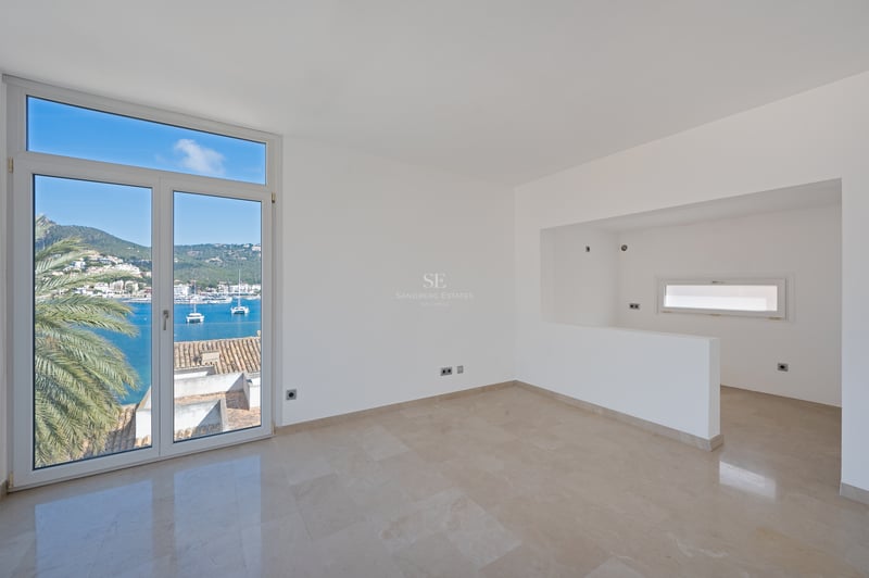 Pièce minimaliste vide avec sols en marbre crème et grandes fenêtres donnant sur une marina méditerranéenne et les montagnes.