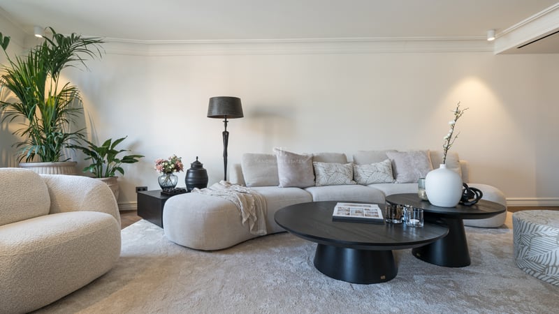 Modern living room with a curved beige sofa, black round coffee tables, and a plush rug in a bright neutral room.