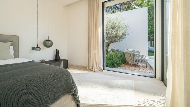 Chambre moderne avec un grand lit, des suspensions design et des baies vitrées ouvrant sur un jardin avec un olivier.