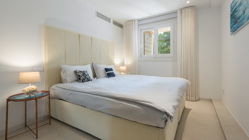 Chambre lumineuse avec tête de lit capitonnée crème, draps blancs et lampes de chevet modernes près d'une fenêtre.