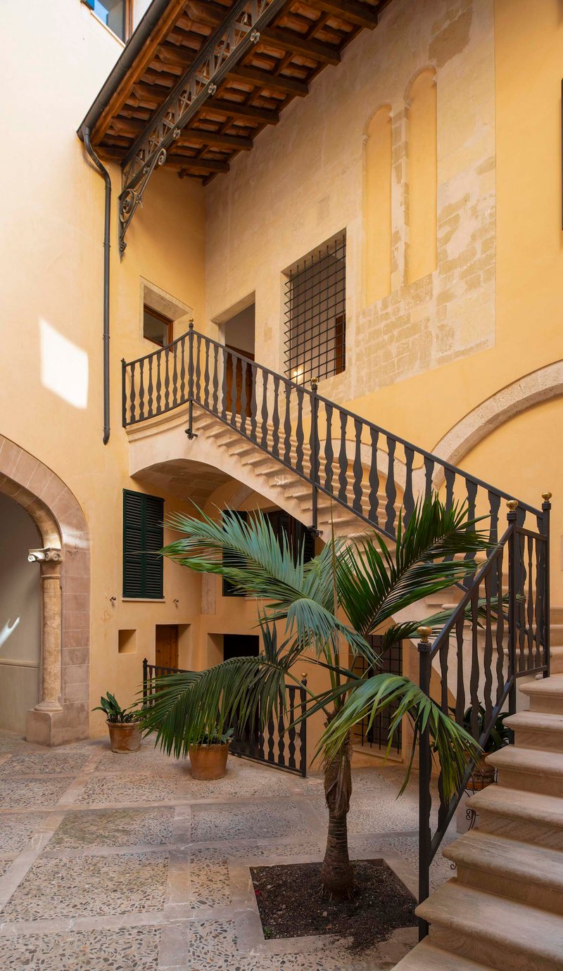 Cour traditionnelle aux murs jaunes, escalier en pierre avec rampe en fer et un palmier central.