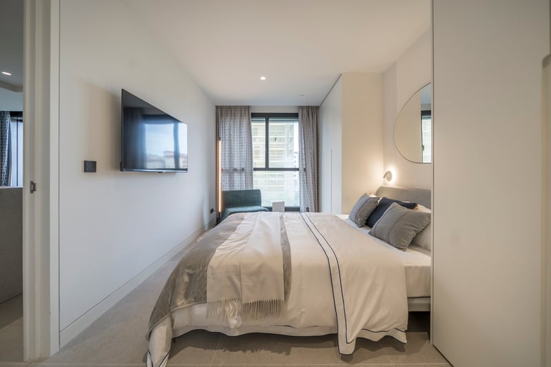 Chambre lumineuse avec TV murale, lit tapissé gris, miroir organique et grande fenêtre.