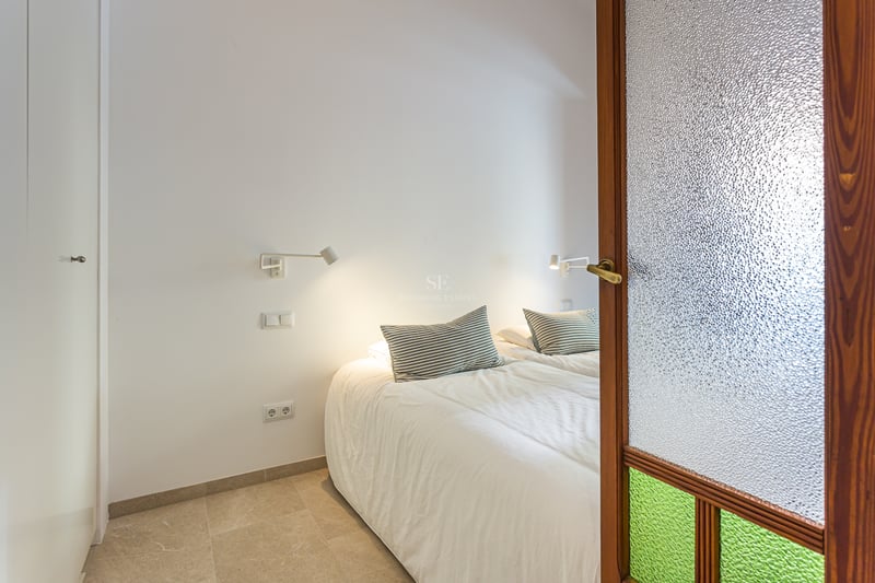 Vue d'une chambre moderne à travers une porte en bois avec verre texturé, lit blanc et liseuses murales.