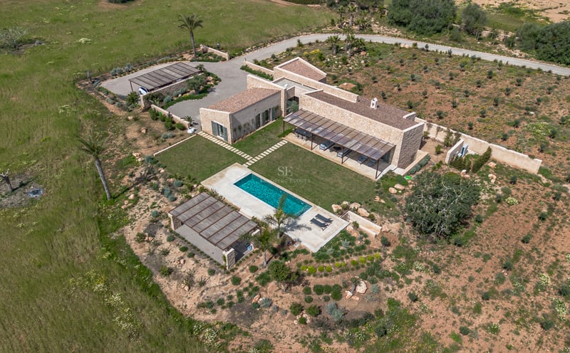 Vista aérea de una villa moderna de piedra con piscina turquesa, jardín de césped y entorno natural en Mallorca.
