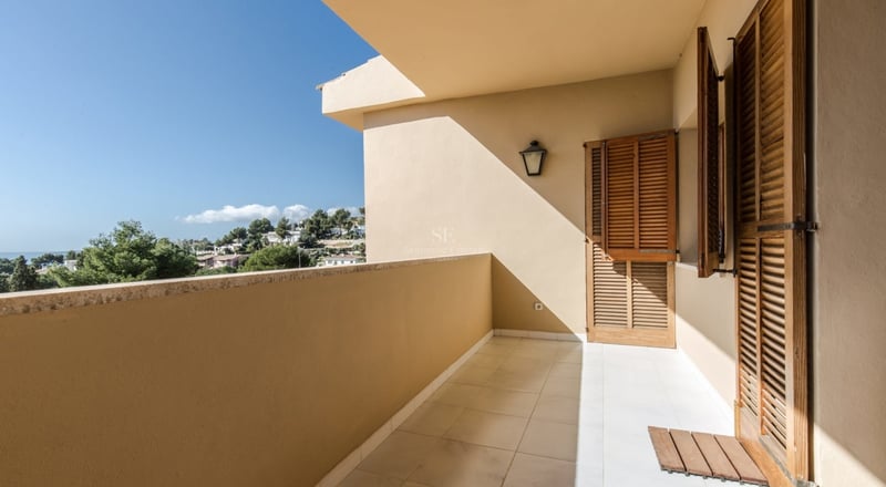 Balcon couvert avec volets en bois et carrelage offrant une vue sur la mer et la verdure sous un ciel bleu.