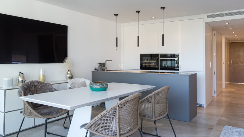 Cocina moderna con isla gris, armarios blancos sin tiradores y zona de comedor con suelo de piedra natural.