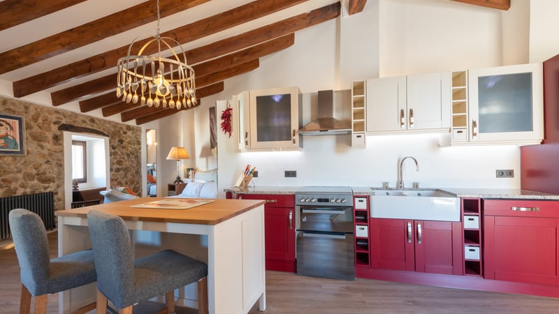 Cuisine avec placards rouges, poutres en bois apparentes et mur en pierre naturelle avec îlot central.