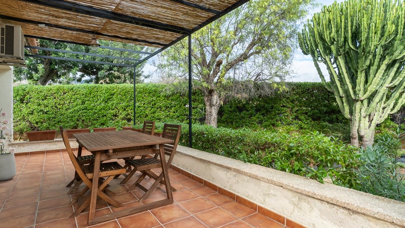 Table à manger en bois sous une pergola en canisse sur une terrasse en terre cuite entourée de verdure.