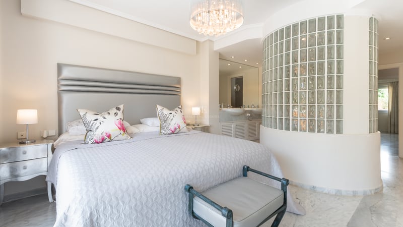 Chambre de luxe avec grand lit, sol en marbre, lustre en cristal et mur incurvé en briques de verre vers la salle de bain.