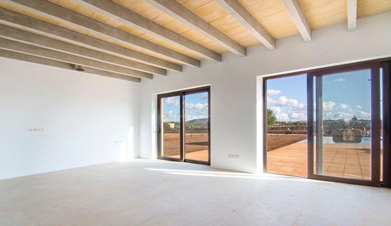 Grande pièce vide avec poutres apparentes en béton et bois, murs blancs et baies vitrées coulissantes.