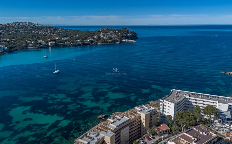Vue aérienne d'une baie turquoise avec des voiliers à l'ancre et des bâtiments côtiers sous un ciel bleu.