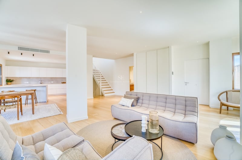 Modern open-plan living room featuring tufted grey sofas, light oak flooring, and a kitchen in the background.