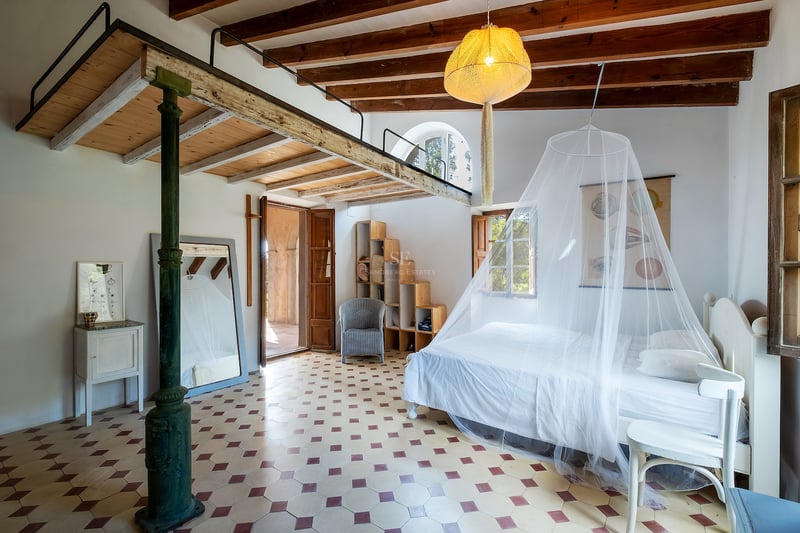 Grande chambre avec lit blanc, moustiquaire, mezzanine en bois, poutres apparentes et sol en carreaux décoratifs.