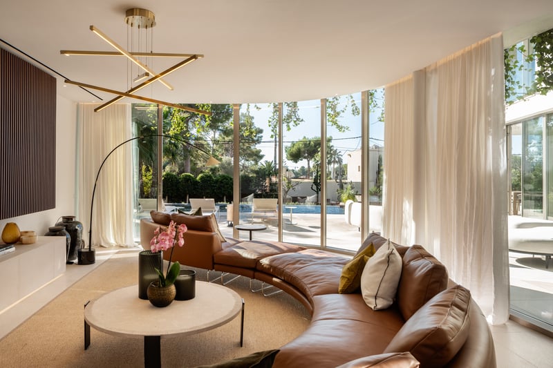Salon moderne avec canapé en cuir courbé, luminaire en laiton et grandes baies vitrées donnant sur une piscine.