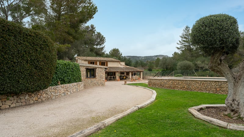 Maison en pierre rustique avec allée en gravier, pelouse verte et piscine en pierre surélevée sous un ciel clair.
