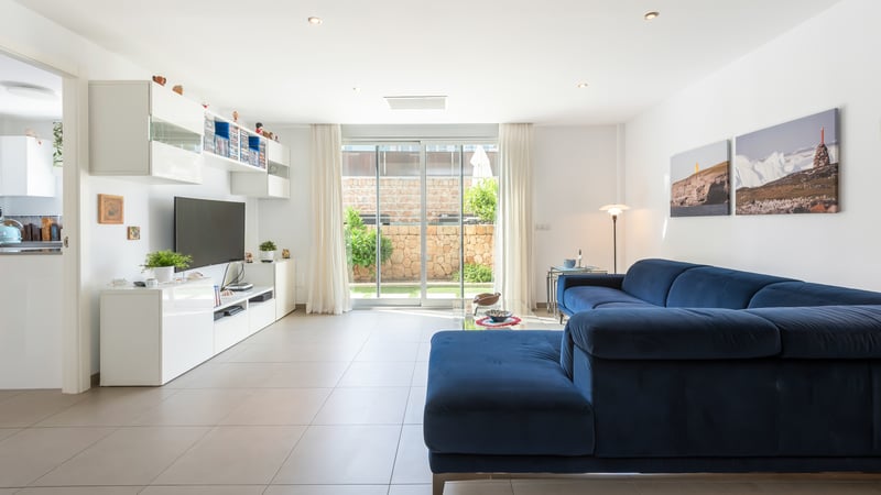 Salon lumineux avec canapé en velours bleu, meuble TV blanc et baies vitrées ouvrant sur un jardin clos de murs en pierre.