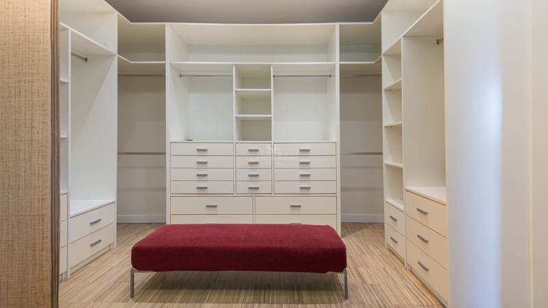 Dressing blanc spacieux avec de nombreuses étagères, tiroirs, sol en bois et un pouf en velours rouge au centre.