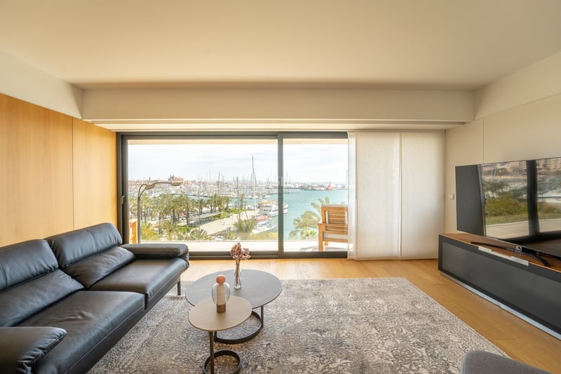Salon contemporain avec canapé en cuir noir et grande baie vitrée offrant une vue sur un port de plaisance.