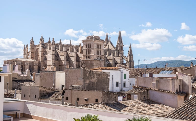 Une grande cathédrale gothique surplombe les toits en terre cuite d'une ville méditerranéenne sous un ciel bleu.