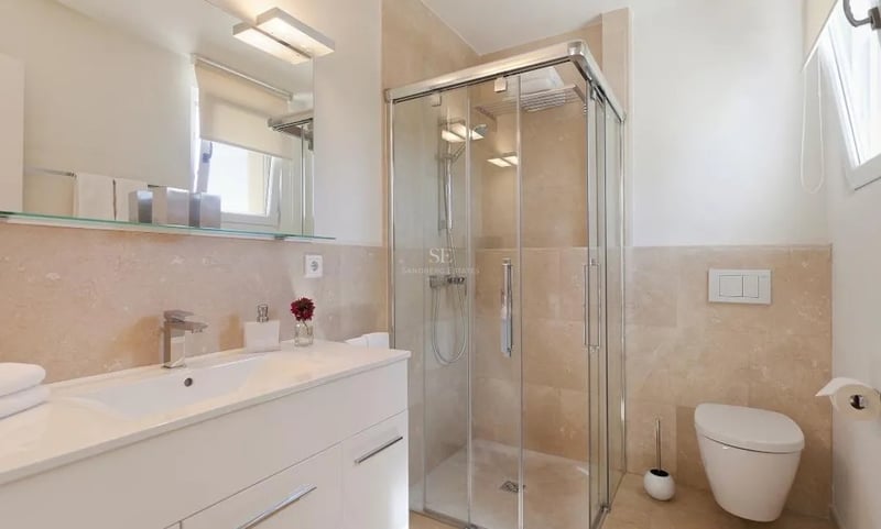 Salle de bain moderne avec douche vitrée, carrelage en marbre crème, meuble vasque blanc et WC suspendu.