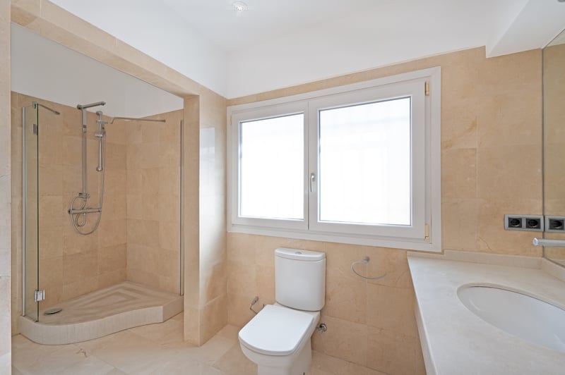 Salle de bain moderne avec murs en marbre crème, douche à l'italienne et grande fenêtre dépolie.
