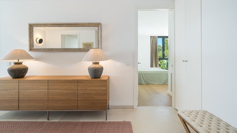 Un buffet en bois avec deux lampes et un miroir orné dans un couloir lumineux menant à une chambre.