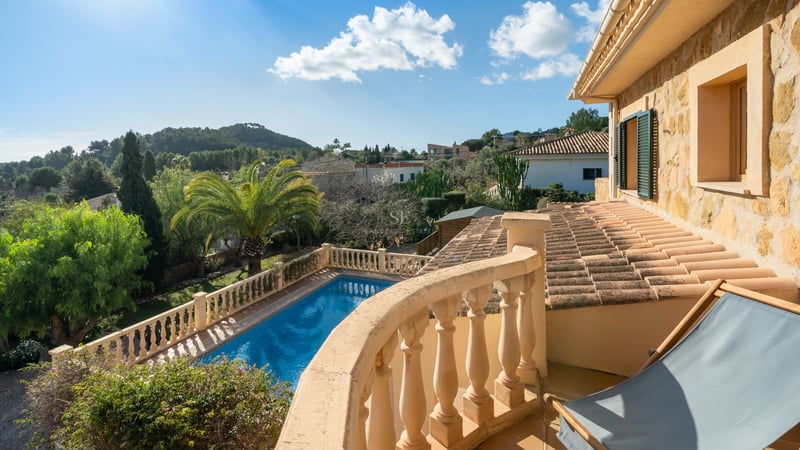 Vue d'un balcon en pierre sur une piscine bleue, des jardins luxuriants et des collines sous un ciel dégagé.