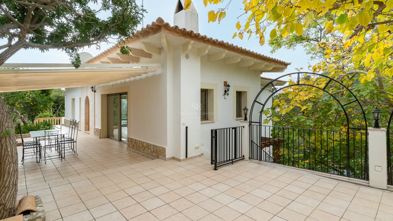 Grande terrasse carrelée avec mobilier en fer forgé, façade blanche et végétation luxuriante sous un auvent.