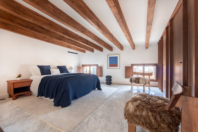 Dormitorio de lujo con vigas de madera oscura, suelo de mármol blanco, ropa de cama azul marino y taburetes de piel.