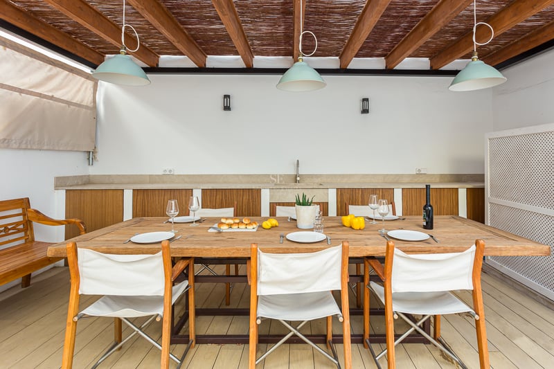 Terrasse couverte avec grande table en bois, chaises en toile blanche et cuisine extérieure sous une pergola.