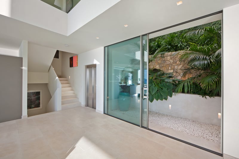 Hall minimaliste avec sol en pierre claire, ascenseur, escalier blanc et baie vitrée donnant sur un patio tropical.