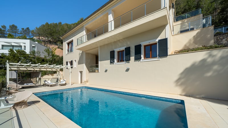Piscine de luxe à côté d'une villa de couleur crème avec balcons en verre et forêt de pins en arrière-plan.