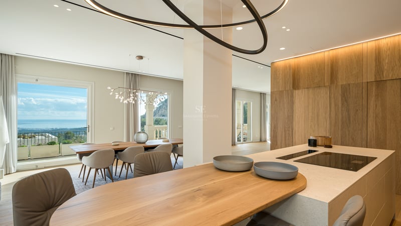 Îlot de cuisine moderne avec plan de travail en pierre et bar en chêne donnant sur la salle à manger et la mer.